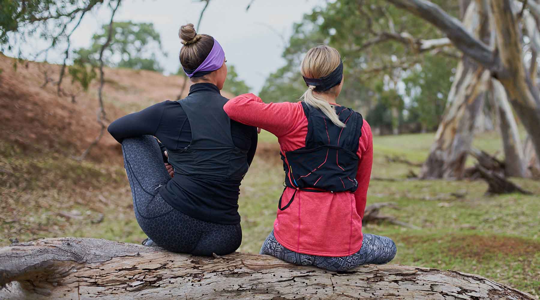 Two women resting on a log with running vests on wearing a purple sweatband and a black headband