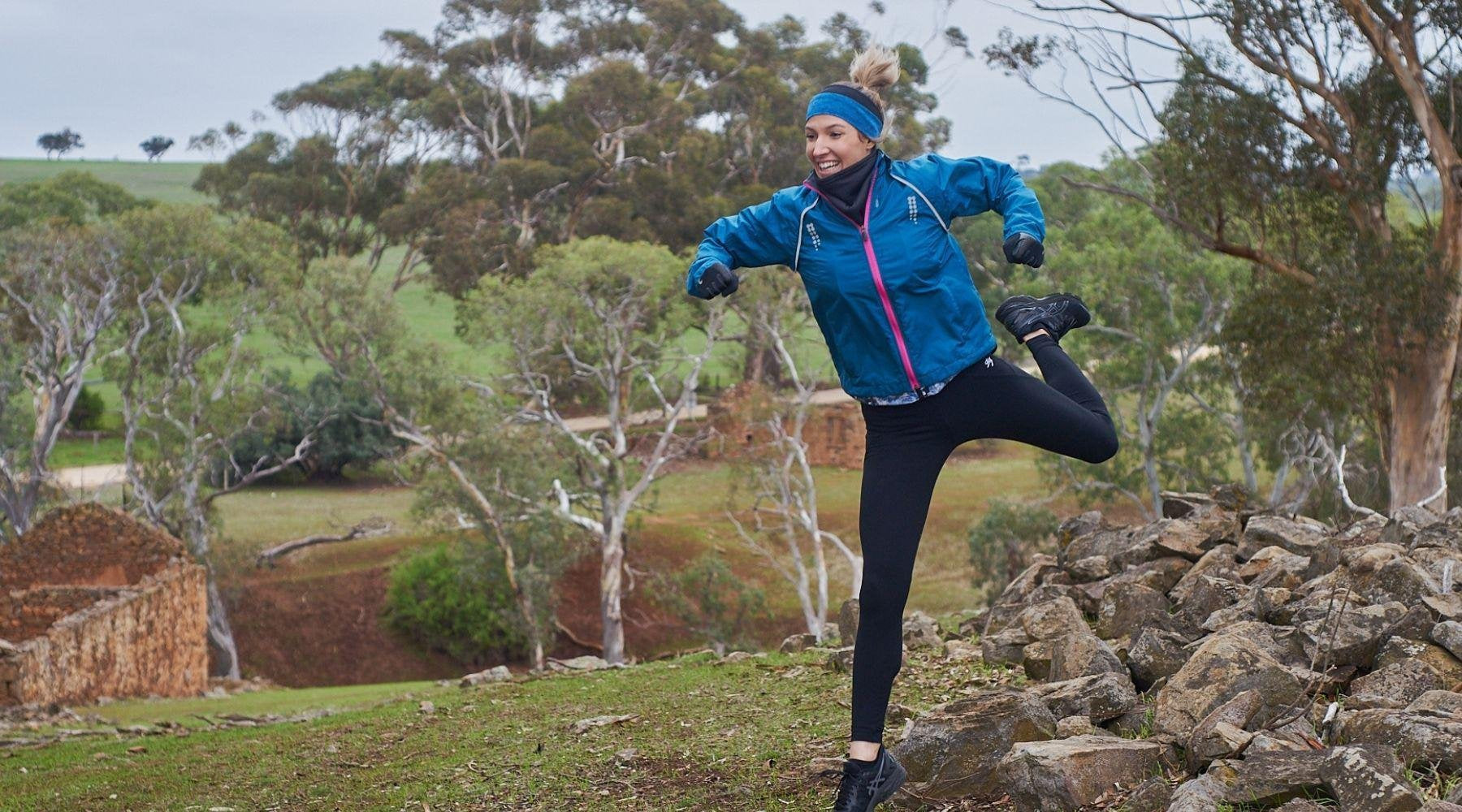 lady jumping off rocks with leg bent in the air with a big smile, wearing polartec neck warmer and reversible winter headband.