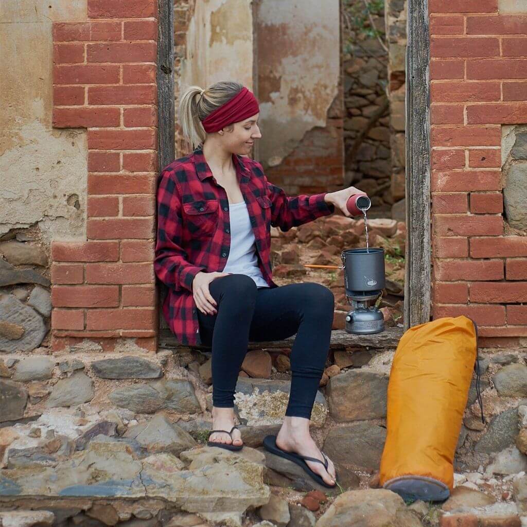 Women siting outside in a ruin pouring water into her hiking water pot wearing red checked shirt and a red bamboo headband